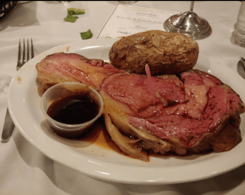 A plate of prime rib with a baked potato and a small cup of sauce, garnished with a pink pick.