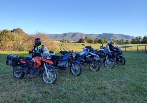 A group of motorcyclists with their bikes parked in a grassy field, mountains visible in the background under a clear sky.