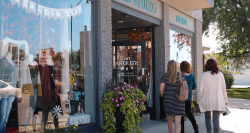 Two women walk past a boutique storefront with colorful flowers and autumn decorations.