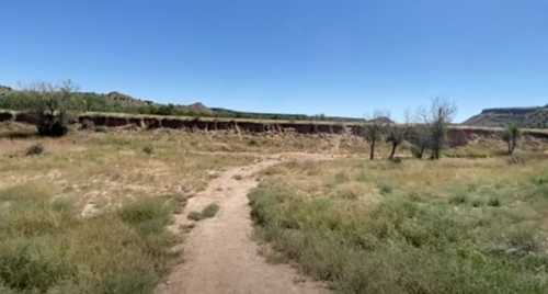 A dirt path leads through grassy terrain with sparse trees and a steep, eroded cliff under a clear blue sky.