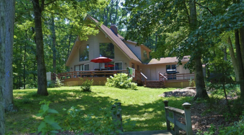 A spacious wooden house surrounded by trees, featuring a deck and a red umbrella, with a small bridge in the foreground.