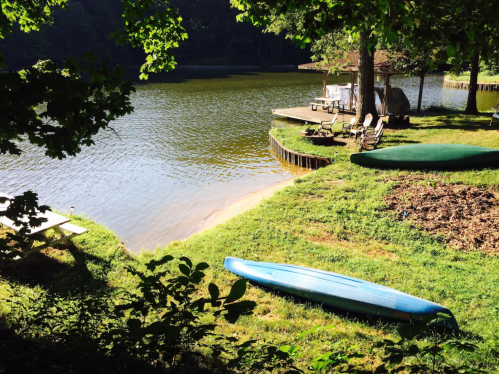 A serene lakeside scene with a blue kayak on the grass, a wooden dock, and chairs by the water under leafy trees.