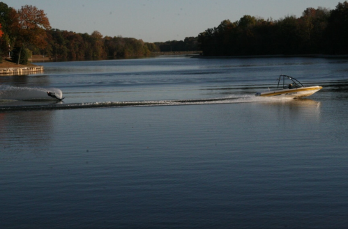 Two boats glide across a calm lake, surrounded by trees with autumn foliage.