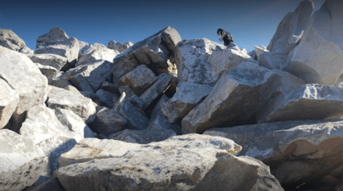 A person climbs a large pile of rocks under a clear blue sky.