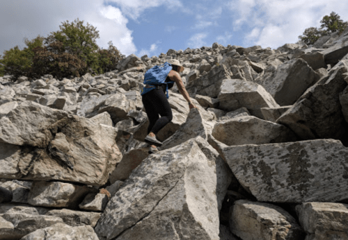 A person climbs a steep rocky slope, navigating large boulders under a partly cloudy sky.