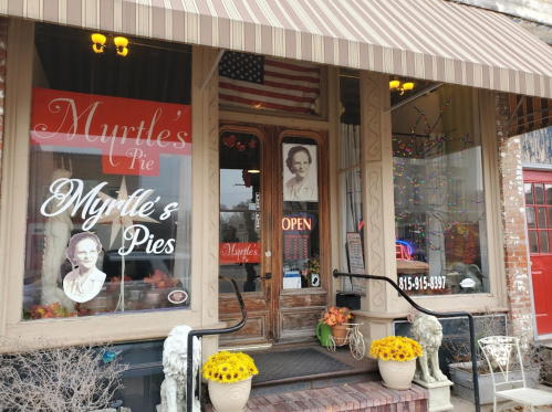 A quaint pie shop with a vintage facade, featuring "Myrtle's Pie" signage, an open flag, and flower pots on the steps.