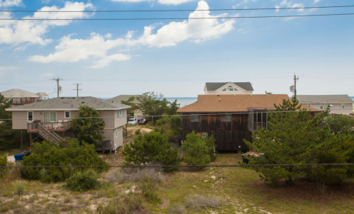 Coastal scene with houses, trees, and a clear sky, overlooking a sandy area near the ocean.