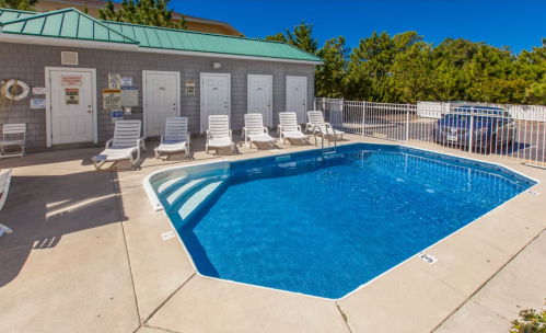 A clear blue pool surrounded by lounge chairs and a building with multiple doors, set in a sunny outdoor area.