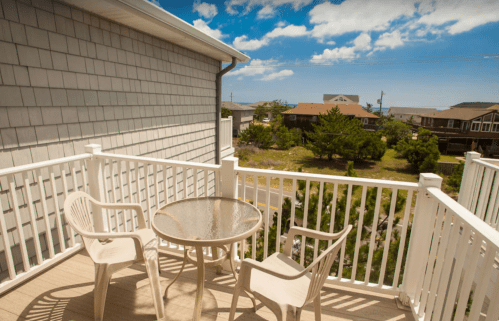 A balcony with two white chairs and a glass table, overlooking a scenic view of houses and trees under a blue sky.