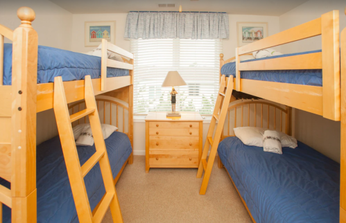 A cozy bedroom with two sets of wooden bunk beds, a nightstand, and a lamp, featuring blue bedding and natural light.