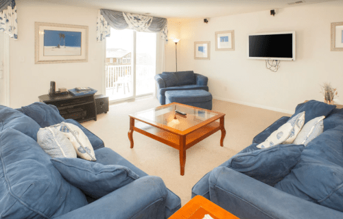 Cozy living room with blue sofas, a wooden coffee table, and a TV, featuring large windows and light-colored walls.