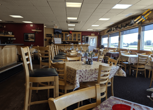 Interior of a restaurant with empty tables, chairs, and a bar area, featuring large windows and a cozy atmosphere.