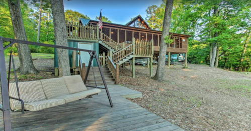 A wooden house surrounded by trees, featuring a porch, stairs, and a swing on a deck in a natural setting.