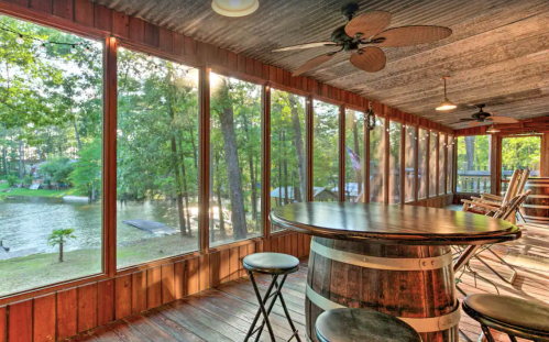 A cozy screened porch with a wooden table, stools, and large windows overlooking a serene lake and trees.