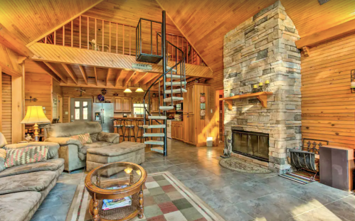 Cozy cabin interior with wooden beams, stone fireplace, spiral staircase, and comfortable seating area.
