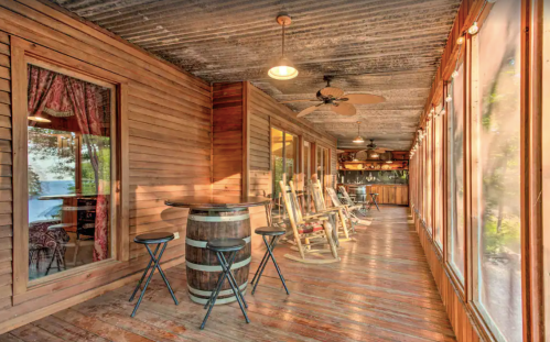 A rustic porch with wooden walls, rocking chairs, a barrel table, and large windows letting in natural light.