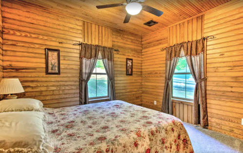 Cozy bedroom with wooden walls, a floral bedspread, and two windows with curtains. A ceiling fan is overhead.