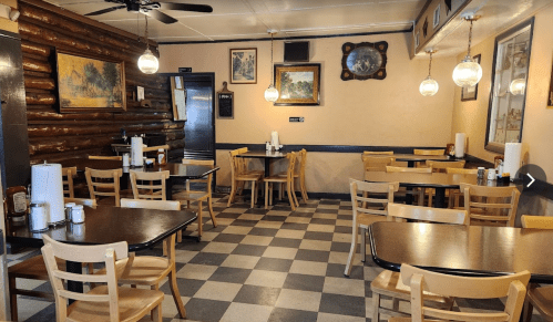 Cozy restaurant interior with wooden walls, checkered floor, and empty tables set for dining. Warm lighting from pendant lamps.