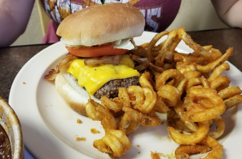 A cheeseburger with lettuce, tomato, and bacon on a bun, served with curly fries on a plate.