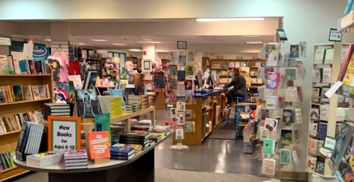 A cozy bookstore interior with shelves of books, a display of new titles, and customers browsing at the counter.
