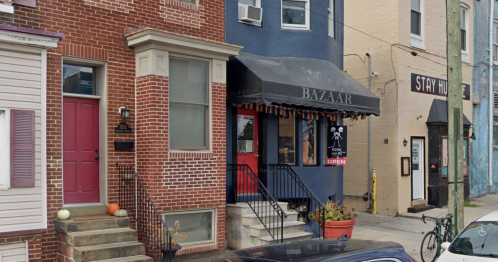 A brick building with a blue facade and an awning labeled "BAZAAR," featuring an open sign and nearby parked cars.