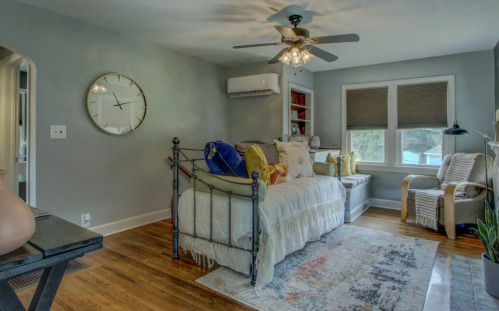 Cozy bedroom with a metal bed, decorative pillows, a ceiling fan, and a window seat, featuring warm wood floors.