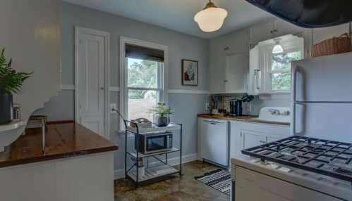 A cozy kitchen with white cabinets, a gas stove, a microwave on a cart, and a window letting in natural light.