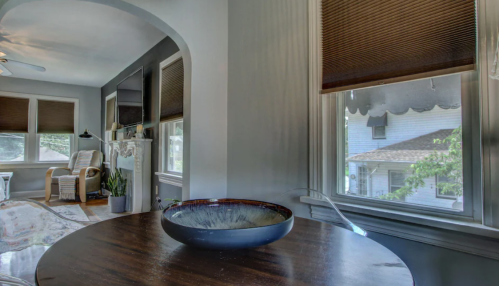 A cozy living room with a wooden table, a decorative bowl, and large windows with blinds, showcasing a bright atmosphere.