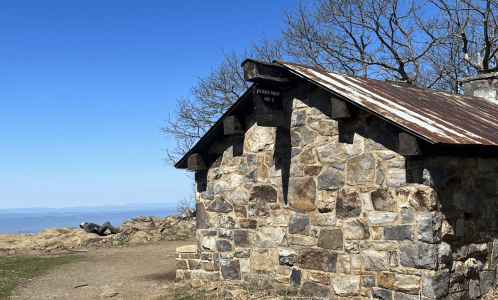 A stone building with a metal roof sits on a rocky landscape under a clear blue sky, surrounded by trees.