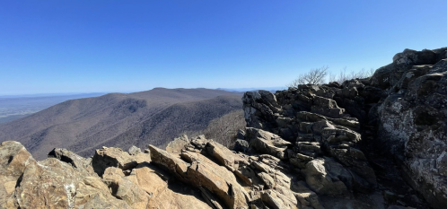 A rocky mountain peak with a clear blue sky and distant hills in the background.