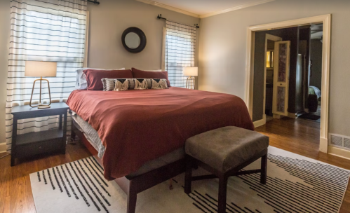 Cozy bedroom featuring a red bedspread, two lamps, a patterned rug, and a view of an adjoining room.
