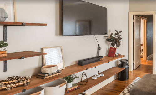 A modern living room with a wall-mounted TV, wooden shelves, decorative items, and a doorway in the background.