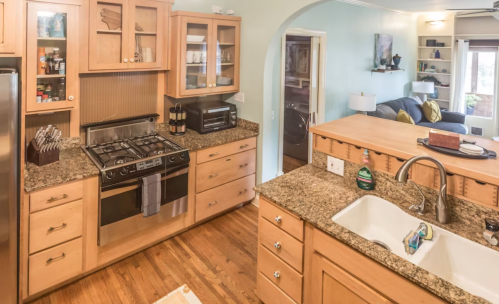 A modern kitchen with wooden cabinets, granite countertops, a gas stove, and an open layout leading to a living area.
