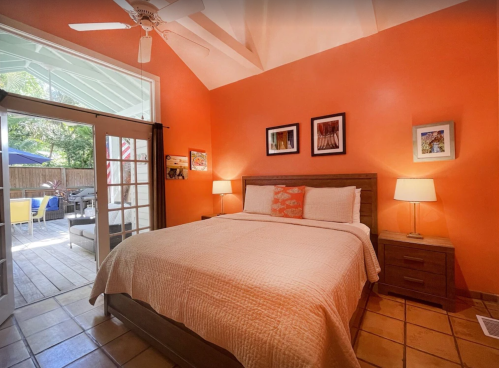 Cozy bedroom with orange walls, a queen bed, nightstands, and doors leading to a patio with outdoor seating.