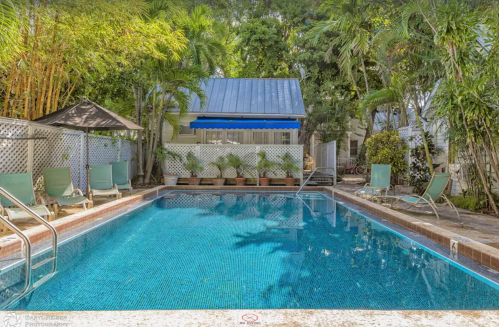 A serene pool surrounded by tropical plants and lounge chairs, with a small building in the background.