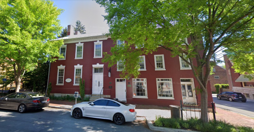 A red brick building with large windows, surrounded by green trees, and parked cars on a quiet street.