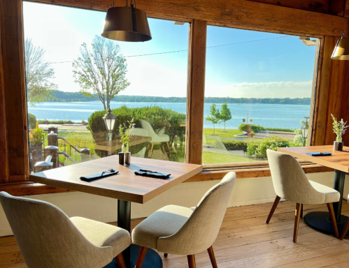 A cozy restaurant interior with two chairs and tables, overlooking a scenic lake through large windows.