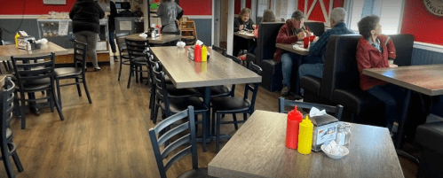 A cozy restaurant interior with wooden tables, black chairs, and patrons enjoying meals. Condiment bottles are on the tables.