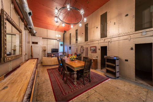 A spacious room with a wooden dining table, chairs, and industrial-style decor, featuring a chandelier and large mirror.