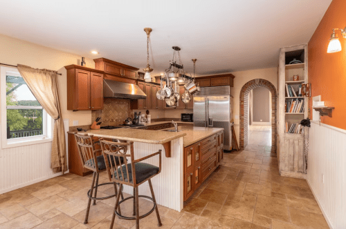 A spacious kitchen with wooden cabinets, a large island, and hanging pots, featuring a warm color scheme and tile flooring.