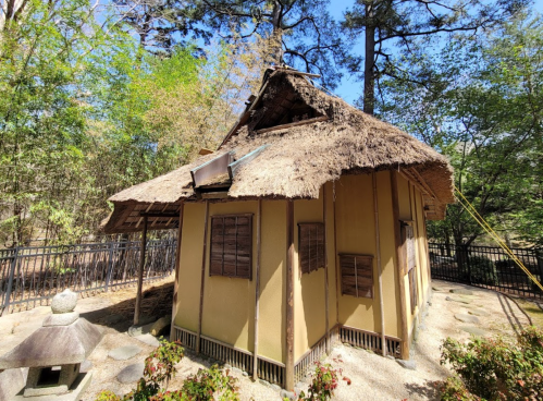 A traditional thatched-roof house surrounded by greenery and a stone lantern in a serene outdoor setting.