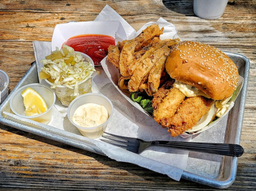 A tray with a sandwich, fried fish, coleslaw, pickles, and dipping sauces, set on a wooden table.