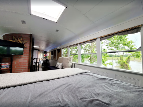 Cozy interior of a converted bus with a bed, large windows, and a view of greenery outside.