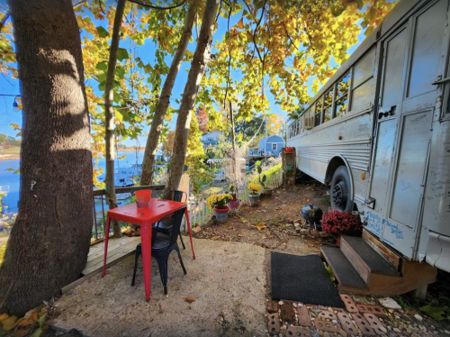 A cozy outdoor space with a red table and chair, surrounded by trees and a vintage bus near a waterfront.
