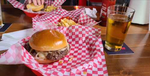 A burger with melted cheese on a bun, fries, and a glass of beer on a wooden table with red checkered paper.