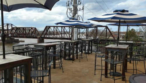 Outdoor seating area with striped umbrellas overlooking a river and a bridge in the background.