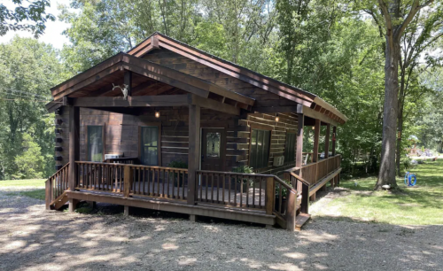 A wooden cabin with a porch, surrounded by trees and gravel, set in a natural outdoor environment.
