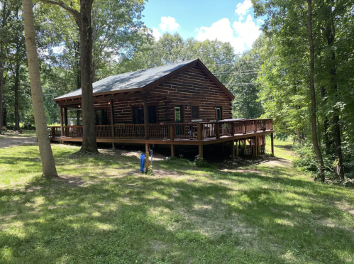 A rustic log cabin surrounded by trees, featuring a spacious deck and a grassy area in front.
