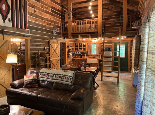 Cozy cabin interior with wooden walls, a leather sofa, and a loft area, featuring rustic decor and warm lighting.