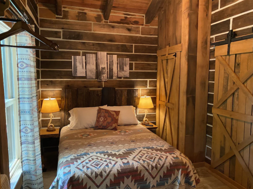 Cozy bedroom with a wooden bed, patterned blanket, two lamps, and rustic log walls. Natural light from a window.